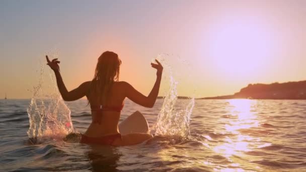 Молодая женщина, сидящая на доске для серфинга, игриво брызгает водой на закате — стоковое видео