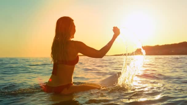 LENS FLARE Молодая женщина сидит на доске для серфинга игриво брызгает водой на закате — стоковое видео