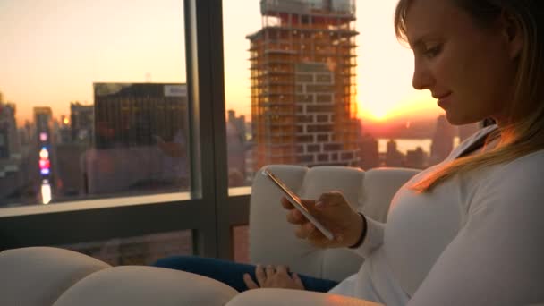 Východ slunce osvětluje New York a dívka textové zprávy, zatímco sedí na gauči — Stock video