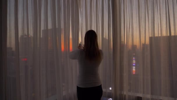FERMER : Femme joyeuse ouvre les rideaux et regarde la ville au coucher du soleil . — Video