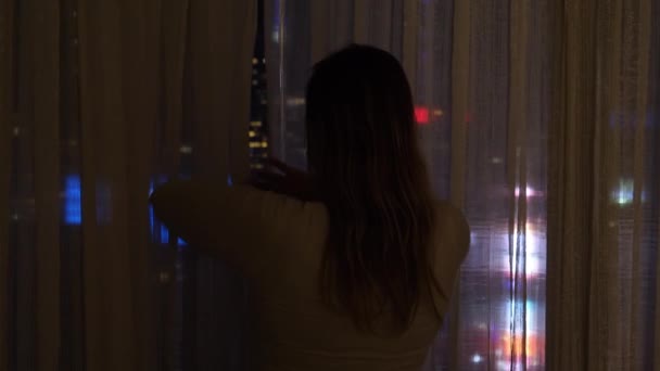 閉じる:女の子はカーテンを開き、ニューヨークの夜の街並みを見ます. — ストック動画