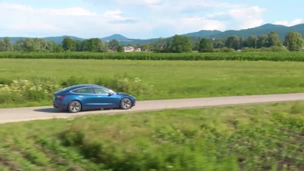 DRONE: Μεταλλικό μπλε Tesla οδηγεί κατά μήκος ενός γραφικού επαρχιακού δρόμου σε μια ηλιόλουστη μέρα — Αρχείο Βίντεο
