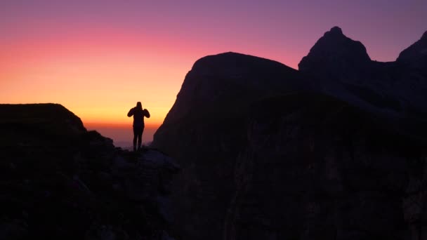 SILHOUETTE: Mulher estende os braços enquanto observa o pôr do sol do topo da montanha — Vídeo de Stock