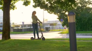 Genç bir kadın, modern şehirde kaldırım boyunca bir e-scooter sürüyor.