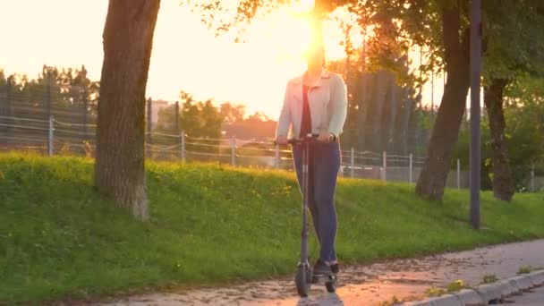 SOL FLARE: Chica alegre sonríe mientras monta un e-scooter a través del parque al atardecer — Vídeos de Stock