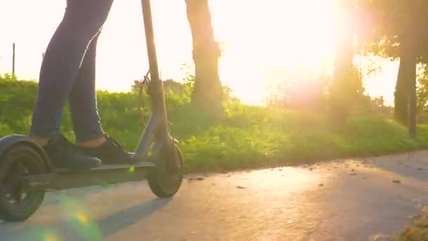 LOW ANGLE: Young woman in jeans rides an electric scooter at beautiful sunrise. — Stock Video