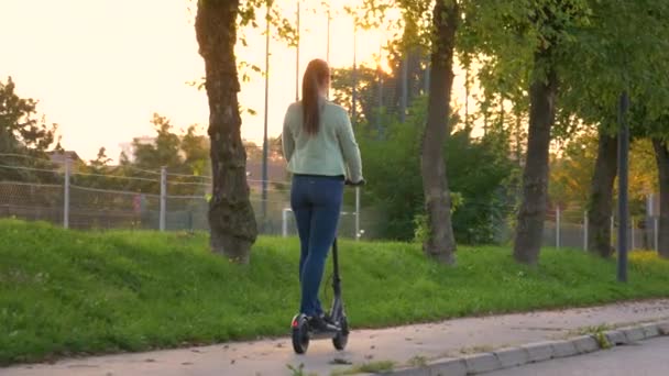 LENS FLARE Mujer monta un scooter eléctrico por una acera vacía en los suburbios — Vídeos de Stock