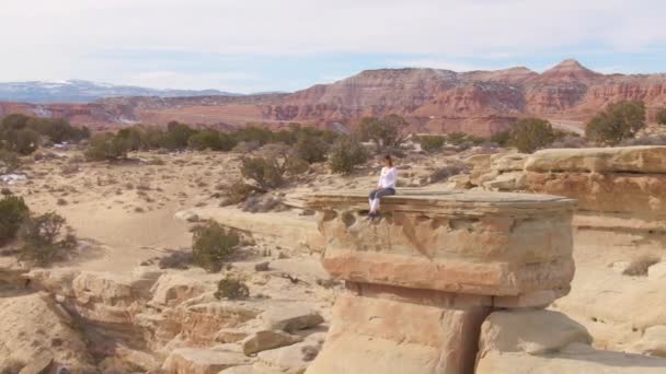 DRONE: Giovane arrampicatore siede sul bordo di una formazione rocciosa che domina il deserto — Video Stock