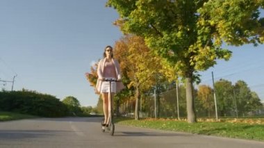 Milenyum kadını elektrikli scooterıyla manzaralı bir caddede yürüyor..