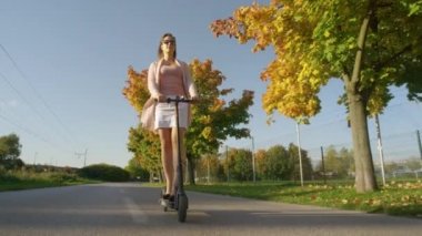 Beyaz etekli, topuklu ayakkabılı, gülümseyen bir kız elektrikli scooter kullanıyor..