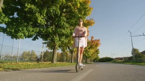 ÁNGULO BAJO: Mujer joven conmuta a lo largo de una avenida de color otoño en un e-scooter. — Vídeo de stock