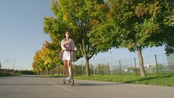 LOW ANGLE: Urban flicka kryssningar längs den natursköna avenyn på en elektrisk skoter. — Stockvideo