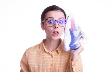 Sarı tişörtlü ve gözlüklü güzel duygusal kız beyaz arka planda bir spor ayakkabısı tutuyor.
