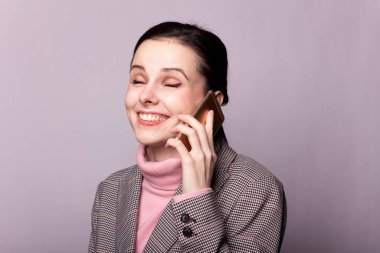 Gri ceketli, pembe balıkçı yakalı güzel bir kız. Gri arka planda telefonla konuşuyor.