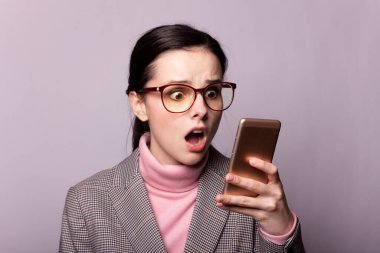 Pembe balıkçı yakalı güzel bir kız, gri ceketli, görüş gözlüklü gri arka planda telefonla iletişim kuruyor.