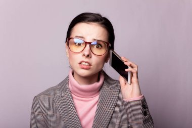 Pembe balıkçı yakalı güzel bir kız, gri ceketli, görüş gözlüklü gri arka planda telefonla iletişim kuruyor.