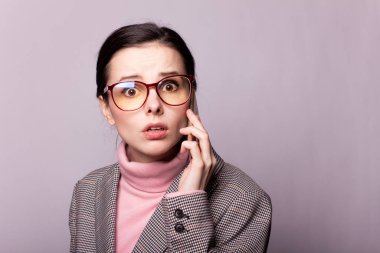 Pembe balıkçı yakalı güzel bir kız, gri ceketli, görüş gözlüklü gri arka planda telefonla iletişim kuruyor.