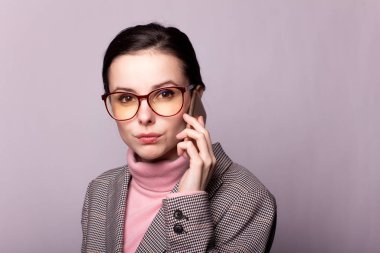 Pembe balıkçı yakalı güzel bir kız, gri ceketli, görüş gözlüklü gri arka planda telefonla iletişim kuruyor.