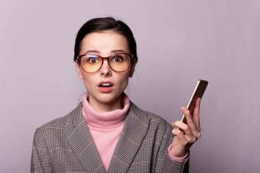 Pembe balıkçı yakalı güzel bir kız, gri ceketli, görüş gözlüklü gri arka planda telefonla iletişim kuruyor.