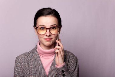 Pembe balıkçı yakalı güzel bir kız, gri ceketli, görüş gözlüklü gri arka planda telefonla iletişim kuruyor.