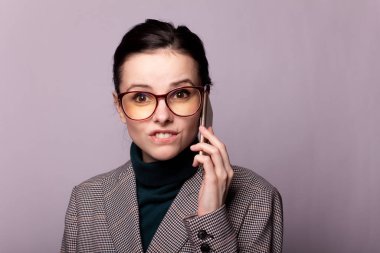 Boğazlı kazaklı, ceketli, gözlüklü gri arka planda telefonla konuşan bir kız.