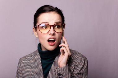 Boğazlı kazaklı, ceketli, gözlüklü gri arka planda telefonla konuşan bir kız.