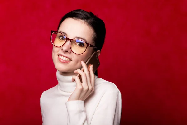 girl in a white turtleneck, glasses for eyesight talking on the phone, portrait on a red background