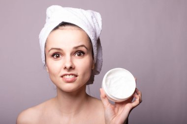 Kafasında beyaz bir havlu ve ellerinde bir kutu krem olan gri arka planı olan bir kız.