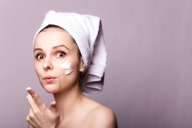 Kafasında beyaz havlu, elinde krem, yüzünde gri arka planı olan bir kız.