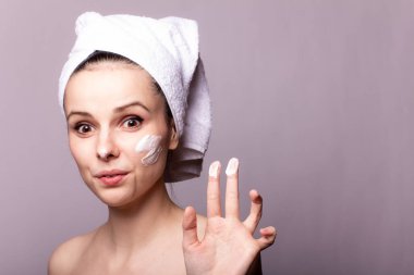 Kafasında beyaz havlu, elinde krem, yüzünde gri arka planı olan bir kız.