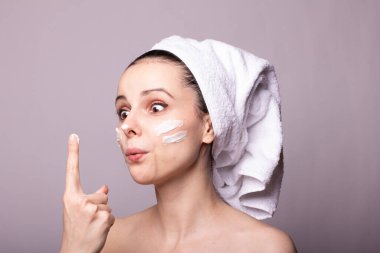 Kafasında beyaz havlu, elinde krem, yüzünde gri arka planı olan bir kız.