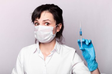 Beyaz gömlekli, tıbbi maskeli ve eldivenli bir kız elinde mavi sıvıyla bir şırınga tutuyor.