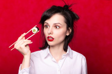 Dudaklarında kırmızı ruj lekesi olan beyaz gömlekli kadın Japon usulü suşi tutuyor.