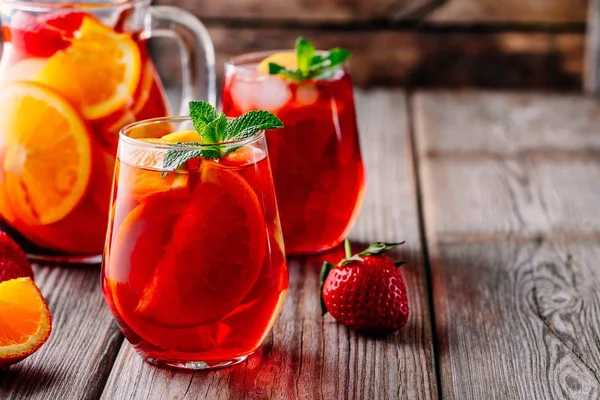 Homemade red wine sangria with orange, apple, strawberry and ice in glass and pitcher on wooden backgroun — Stock Photo, Image