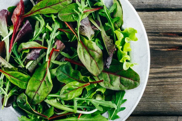 Mix of fresh green salad leaves with arugula, lettuce, spinach and beets on wooden rustic background. — Stock Photo, Image