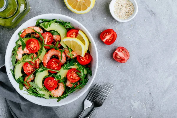 Healthy arugula salad bowl with shrimp, avocado, tomato, and sesame seeds on gray stone background. — Stock Photo, Image