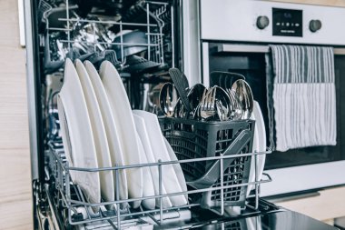 Modern İskandinav mutfağında yıkandıktan sonra beyaz temiz bulaşıklarla bulaşık makinesini aç. Açık bulaşık makinesindeki mutfak malzemelerini temizle..