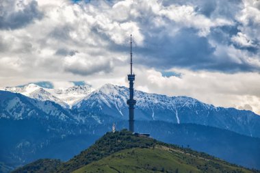 Bulutlu gökyüzü ve dağlardan oluşan bir arka planda televizyon kulesi