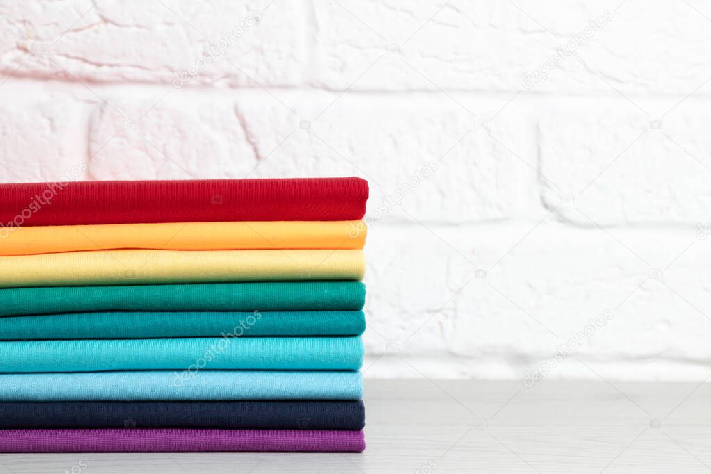 A stack of clean, neatly folded colored clothes on a light wooden table. Colors of rainbow. Place for text