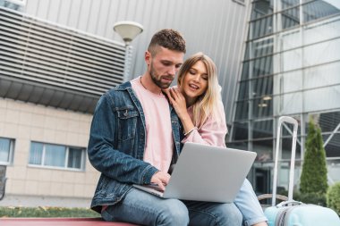 Heyecanlı genç çift havaalanında uçuşu bekliyor. Adam oturuyor ve dizüstü bilgisayar kullanıyor..