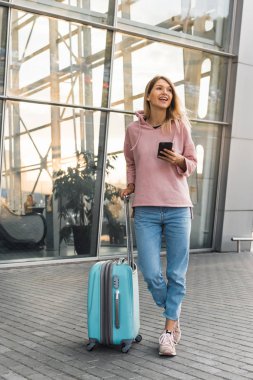 Havalimanında dikilirken akıllı telefon kullanan gündelik giysili genç sarışın kadın turist..