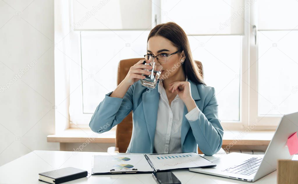 Pretty young woman in the office drinking water while working.