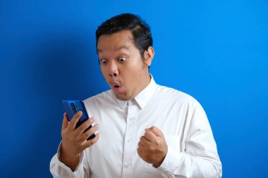 Asyalı erkek ofis çalışanı akıllı telefonuna erişirken kendinden emin görünüyor. Adam şaşırmış bir ifade sergiliyor.