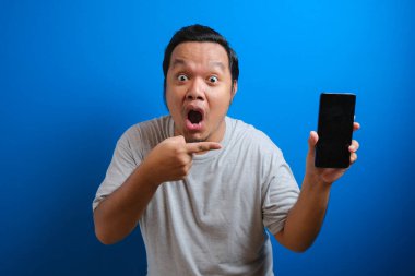 Gri tişörtlü, cep telefonunu işaret eden ve kameraya hayretle bakan, mobil veri tarifelerine şaşırmış şişman Asyalı adamın portresi. Stüdyo çekimi mavi arkaplanda izole edildi