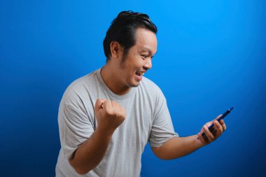 Gri tişört giyen şişman Asyalı adam akıllı telefonundan aldığı iyi haberden memnun görünüyor. Erkekler zafer işaretleri gösterir. mavi arkaplan