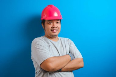 Kırmızı kask takan şişman Asyalı adam kameraya gülümsüyor ve kendinden emin bir hareketle kollarını kavuşturuyor.