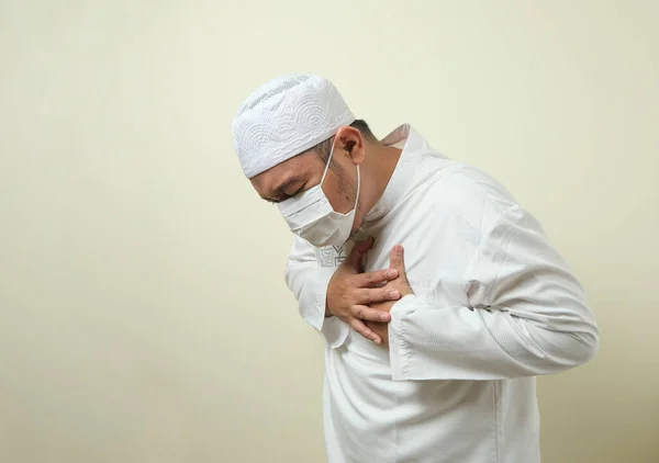 a asian muslim fat guy wearing mask feeling pain in his chest, gesture holding left chest.