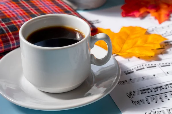 Tasse de café sur une soucoupe sur un fond bleu. notes musicales et une tasse de café avec une serviette à carreaux rouges sur une table bleue. feuilles jaunes et rouges d'automne reposent sur une feuille avec des notes — Photo