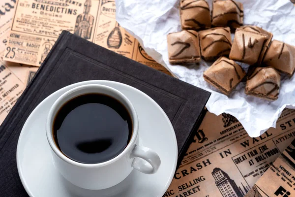 Une tasse de café fort sur une soucoupe blanche et un livre noir sur une table couverte de vieux journaux. tasse avec café et gaufres sur papier blanc froissé. arrière-plan vintage de vieux journaux — Photo