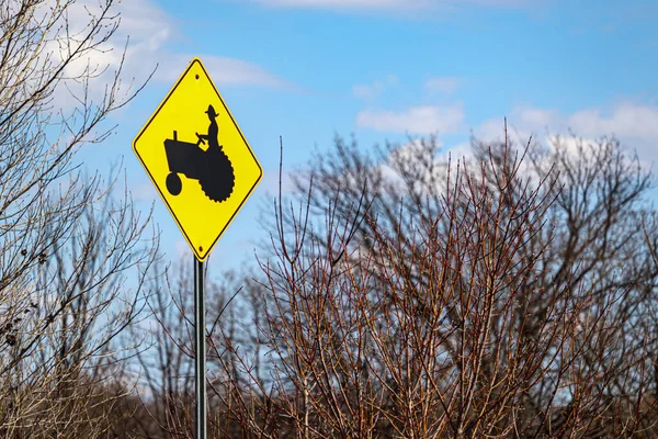 A yellow sign shows a black illustration of a farmer riding on a tractor, marking a common tractor crossing on a road by farmland.